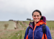 Mary-Ann wears a blue and orange raincoat and smiles at the camera. behind her there's a path that tracks to the horizon. the weather looks wet