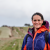 Mary-Ann wears a blue and orange raincoat and smiles at the camera. behind her there's a path that tracks to the horizon. the weather looks wet